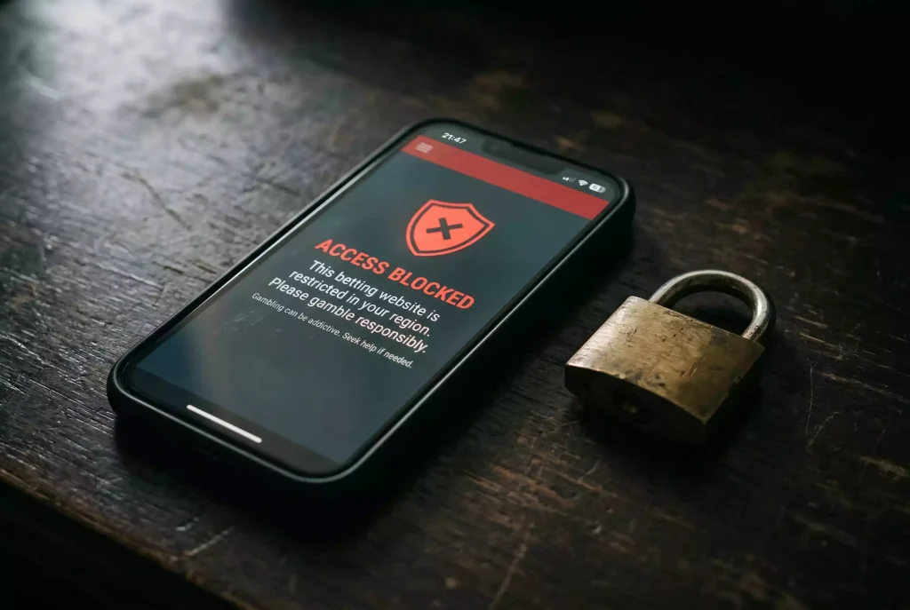 Smartphone mit einer gesperrten Wettseite und einem Warnsymbol auf dem Bildschirm, daneben ein Vorhängeschloss